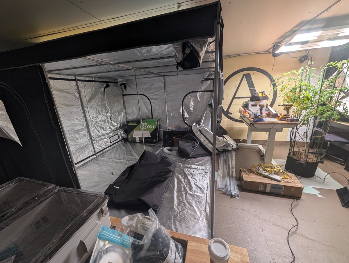 Biodomul real: cortul de creștere argintiu, lampa, o plantă de roșii și masa cu hardware, cu logoul AutonCorp pe perete.